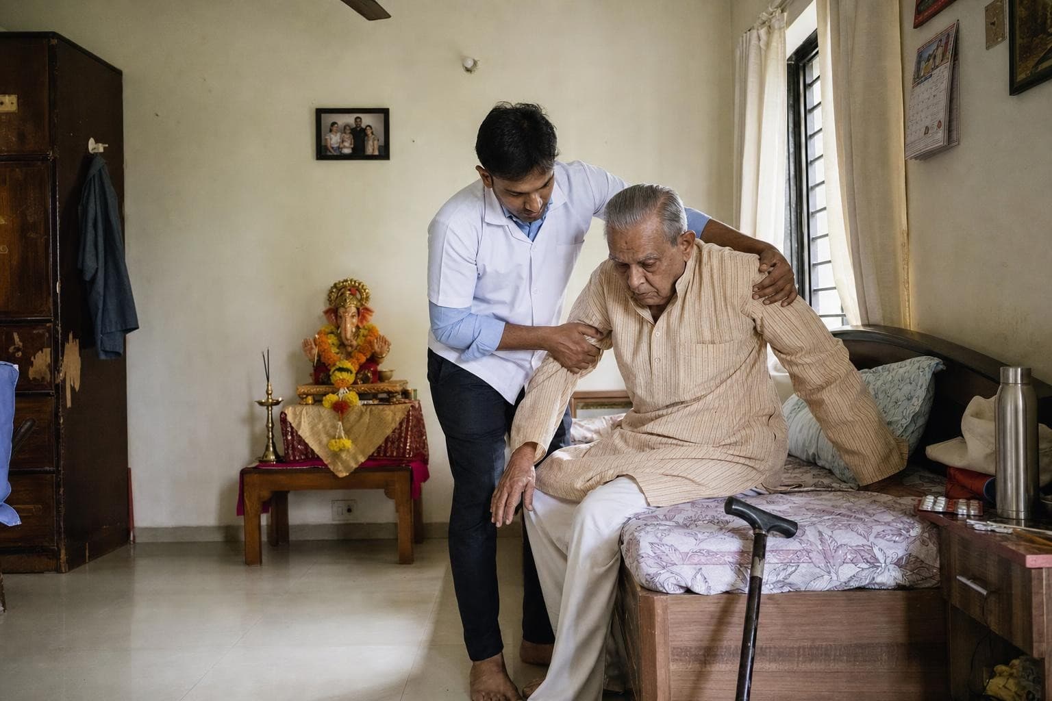 A Healkin nurse attendant helping an elderly man sit up in bed in a Mumbai flat, with a small decorated Ganpati idol on a table behind them
