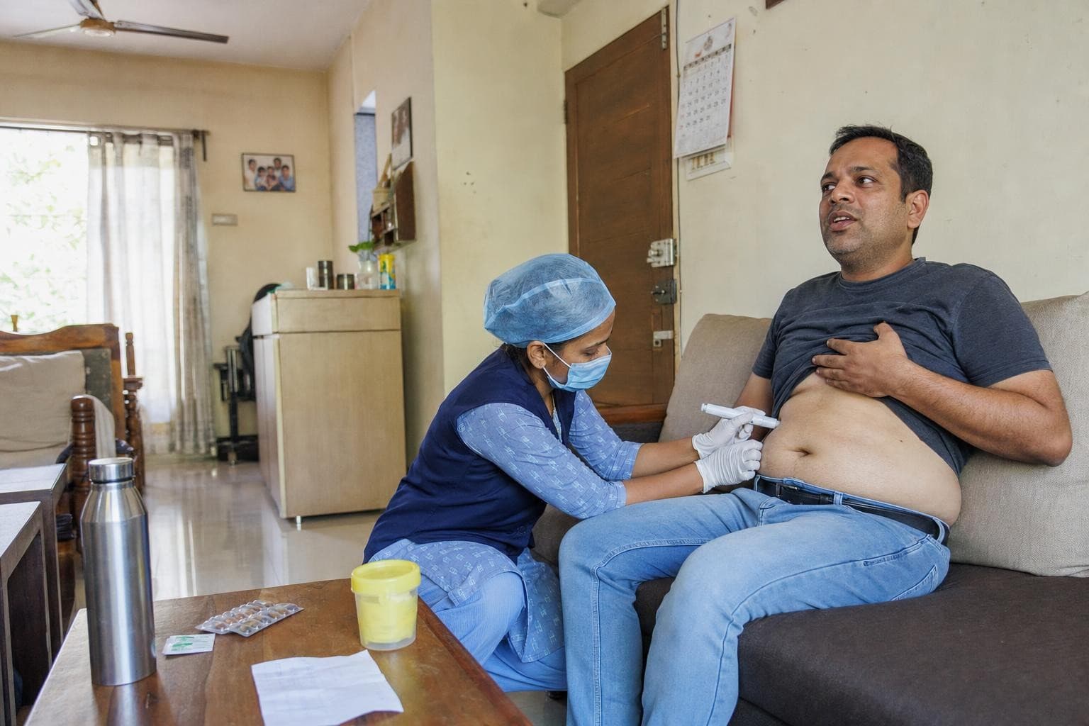 A Healkin nurse giving a subcutaneous injection with a pen device into the abdomen of a man reclining on his sofa at home