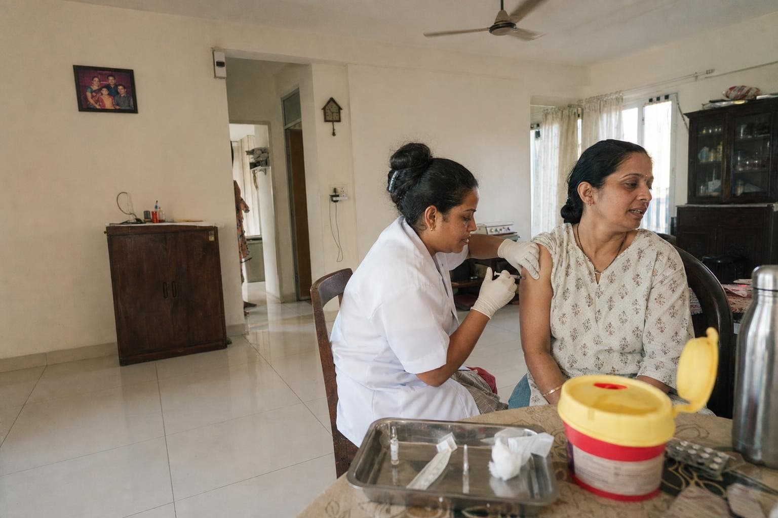 A Healkin nurse giving an intramuscular vitamin injection into the upper arm of a woman seated at her dining table at home